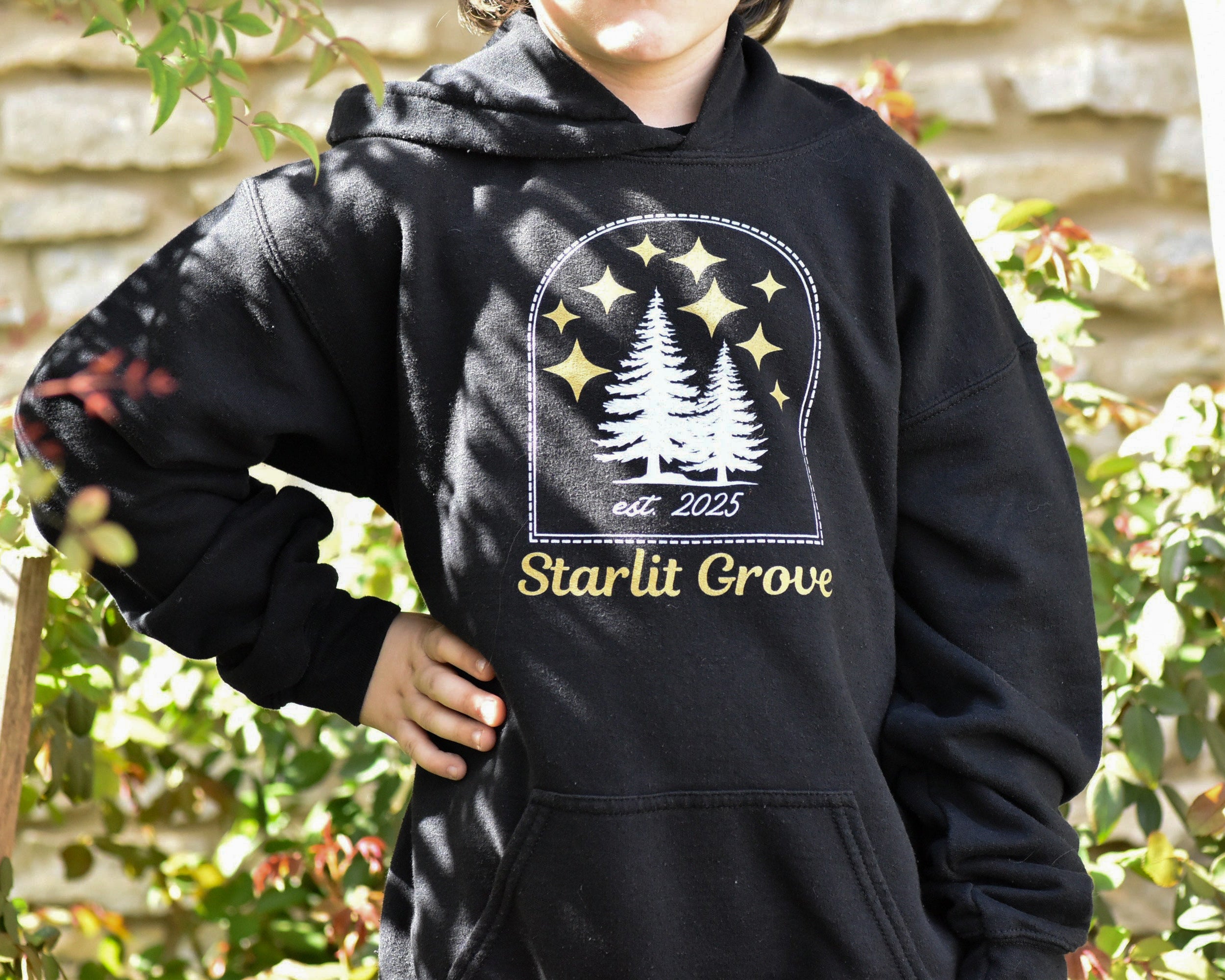 Person wearing a black hoodie with 'Starlit Grove' design outdoors.