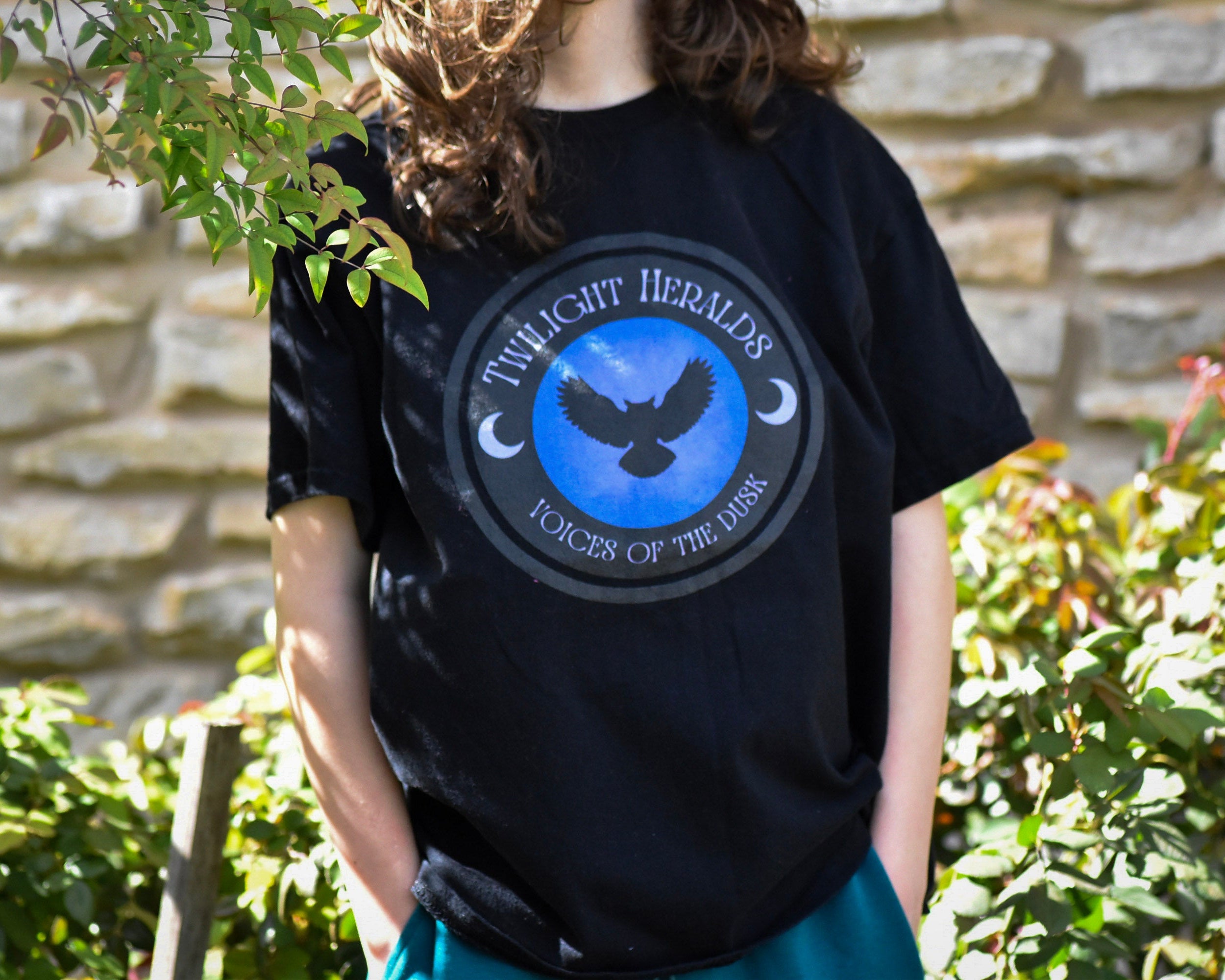 Person wearing a black t-shirt with a blue logo and text, standing outdoors against a stone wall.