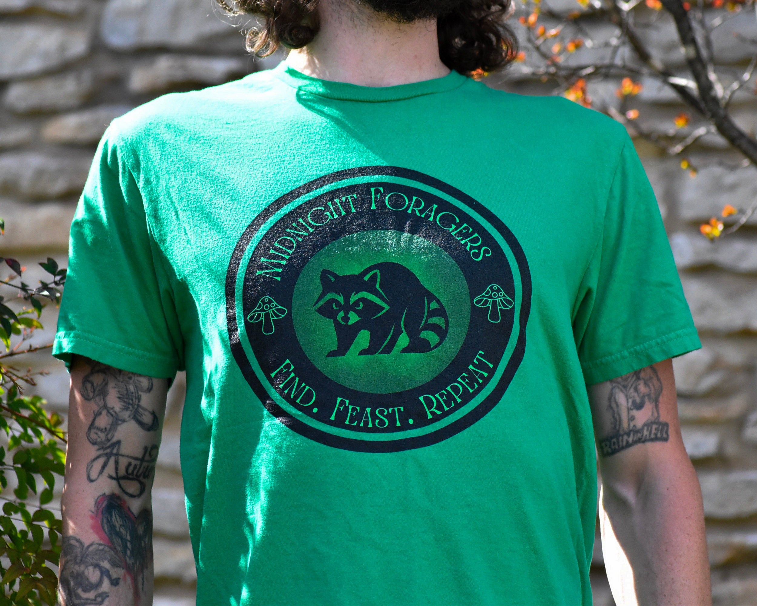 Person wearing a green t-shirt with a raccoon graphic and text, standing against a stone wall.