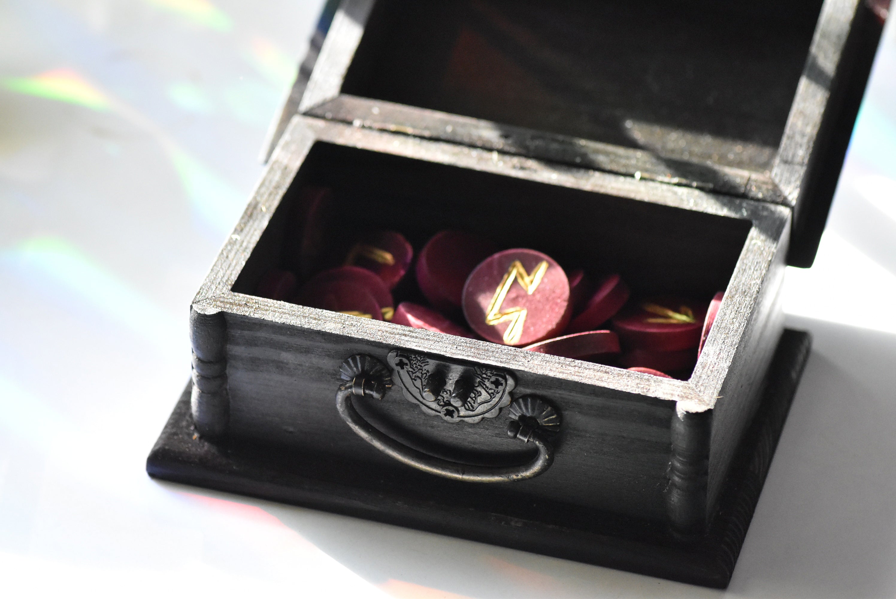 Black jewelry box with a silver handle containing red and gold wooden circle runes on a white background.