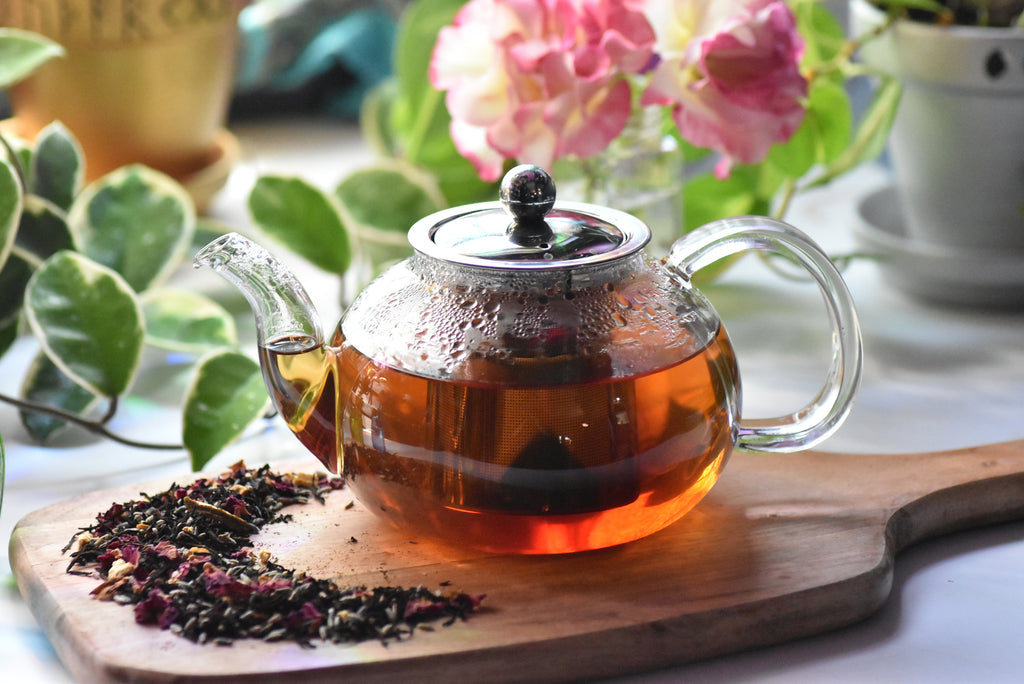 Glass teapot with hot tea on a wooden board, surrounded by dried tea leaves and flowers.