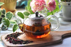 Glass teapot with hot tea on a wooden board, surrounded by dried tea leaves and flowers.