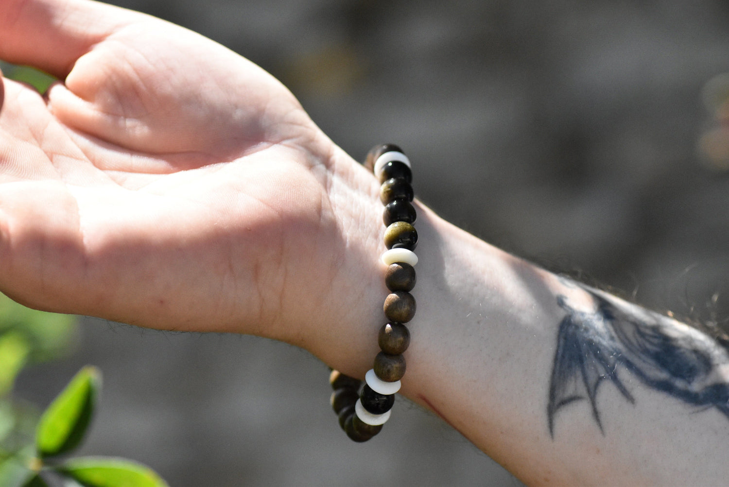 Hand wearing a beaded bracelet with a blurred natural background. The Bracelet has grey wooden beads alternated with golden obsidian. These two colors are divided by thin ox bone beads. 