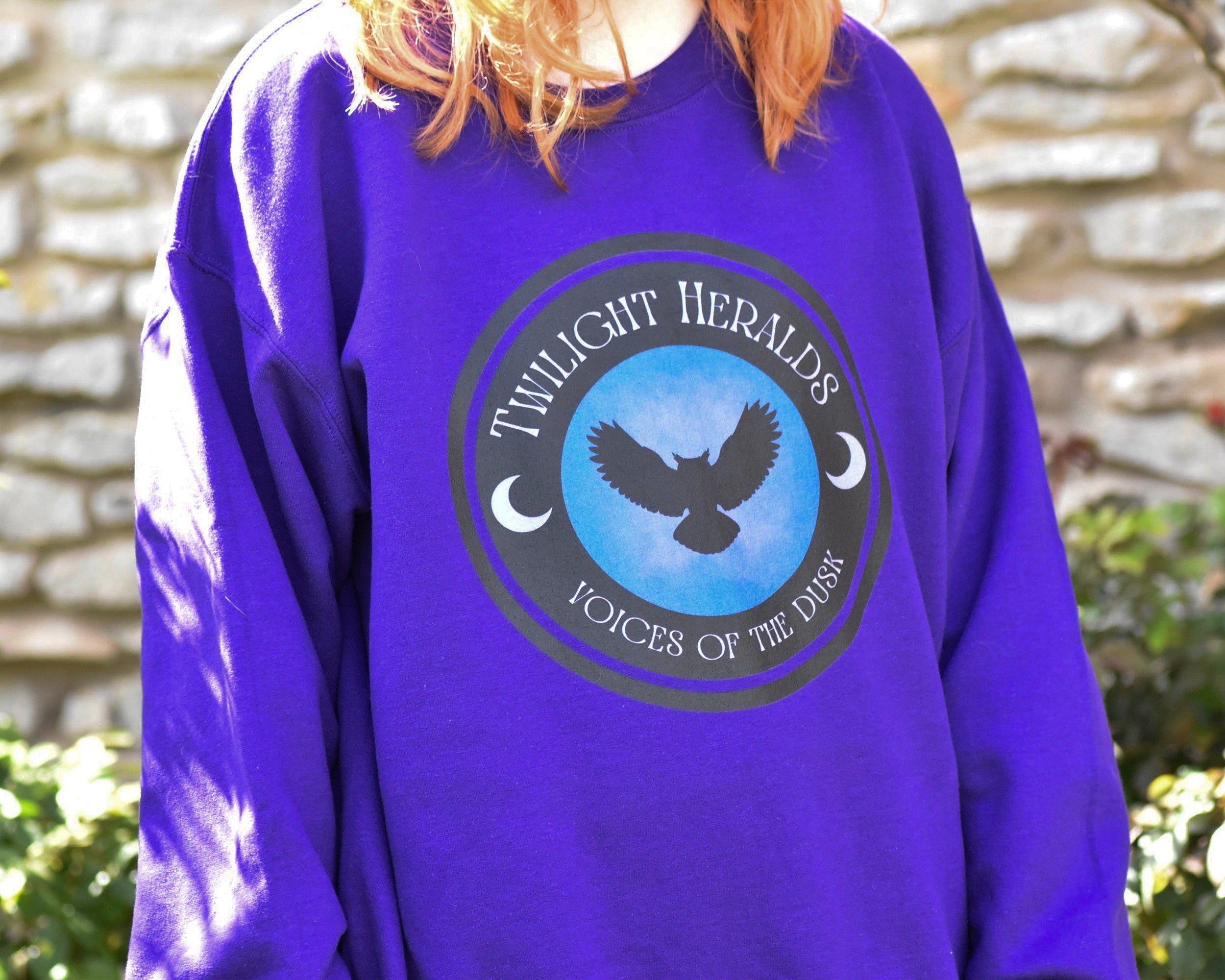 Person wearing a purple sweatshirt with a logo and text against a stone wall background