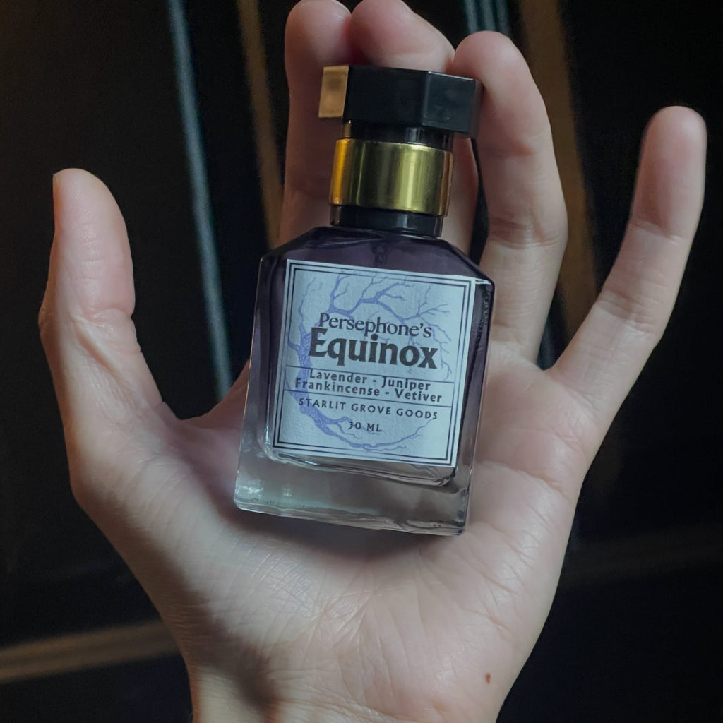 A hand holding a bottle of Persphone's Equinox Eau de Parfum, with a beaded bracelet visible on the wrist against a dark background.
