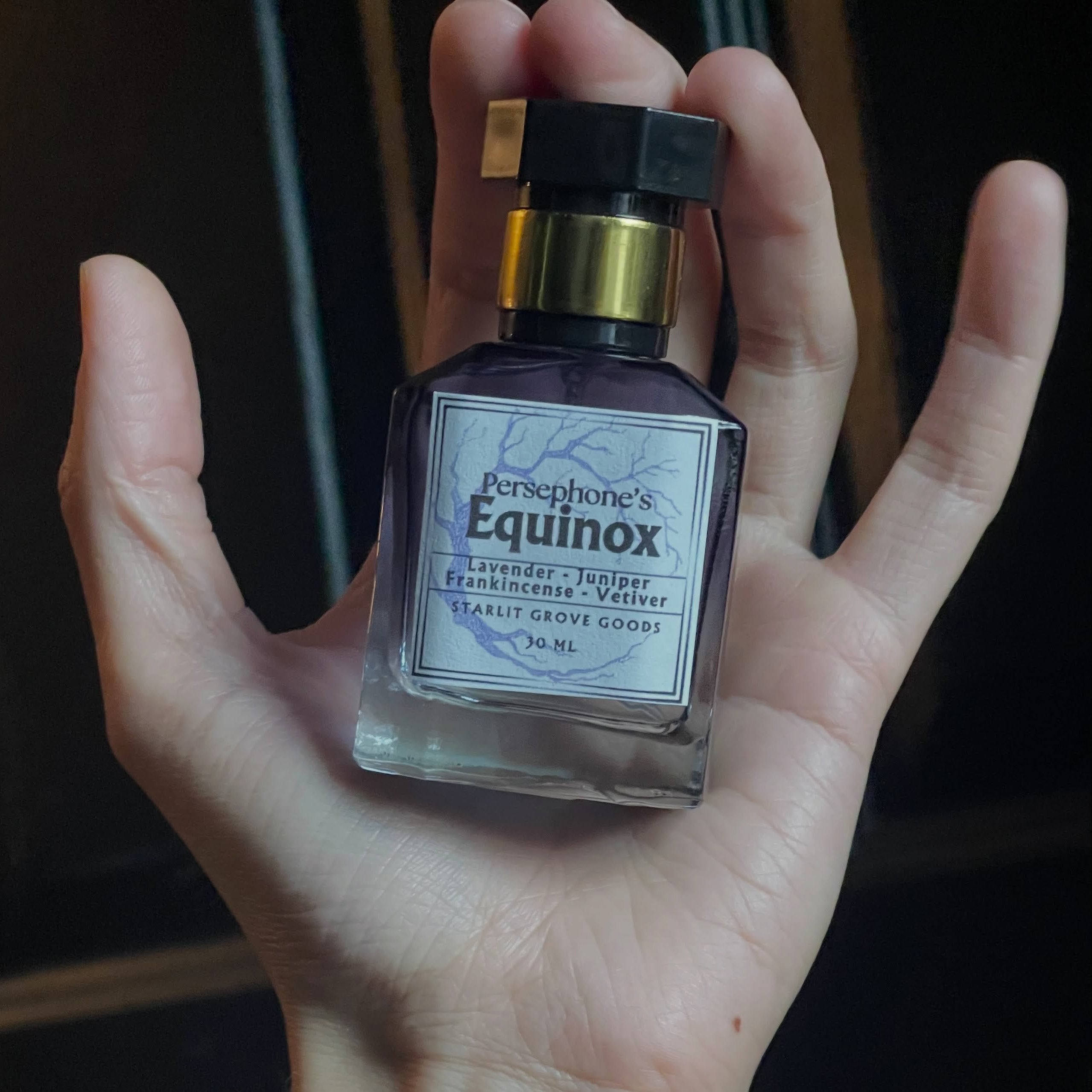 A hand holding a bottle of Persphone's Equinox Eau de Parfum, with a beaded bracelet visible on the wrist against a dark background.