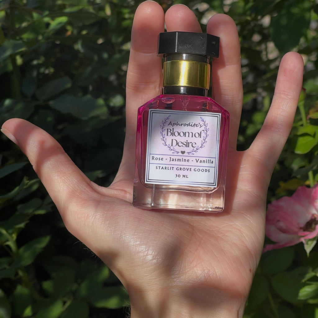 A hand holding a small perfume bottle labeled Aphrodite's Bloom of Desire 